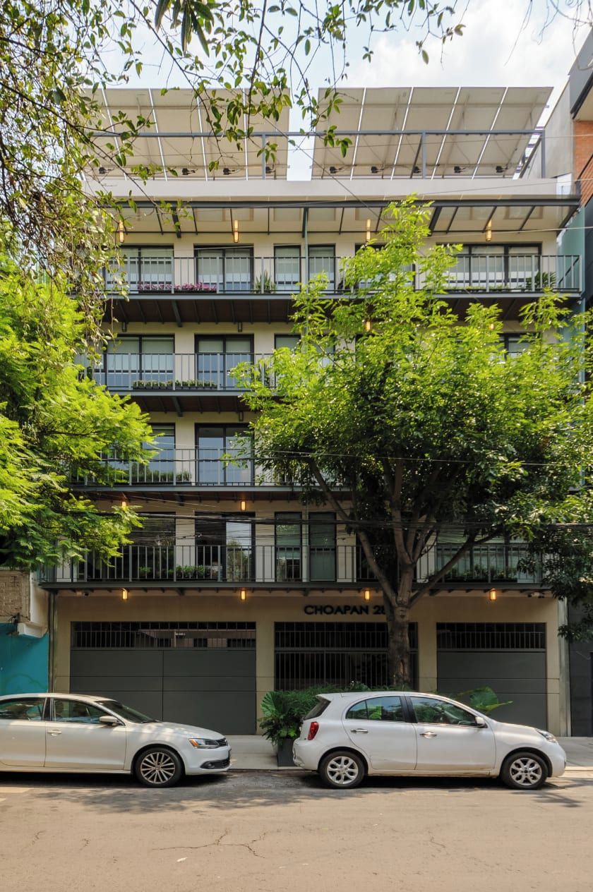 A multi-story building with balconies, a car parked in front, and a tree in the foreground.