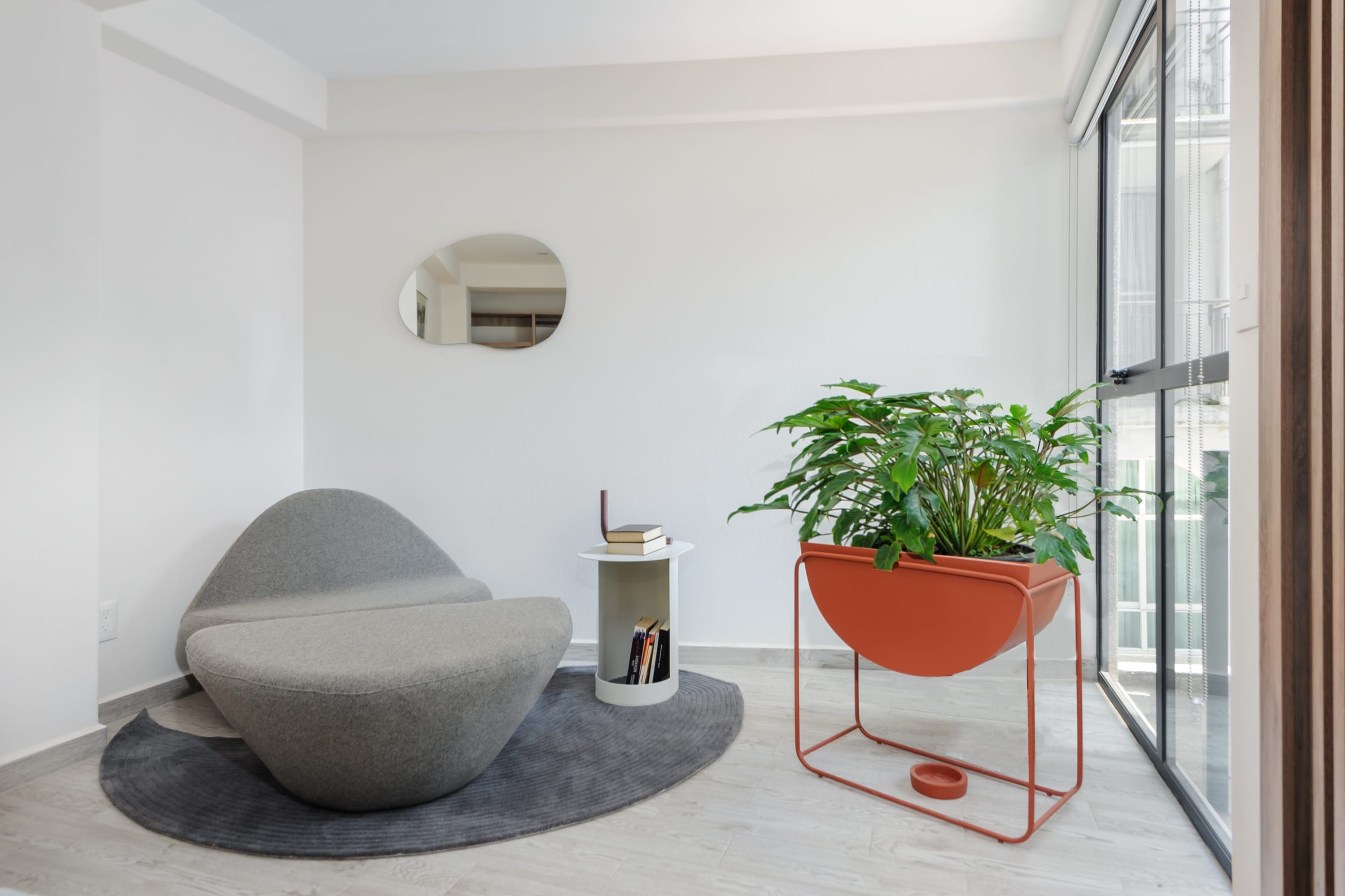 A modern room with a grey bean bag chair, a small white side table with books, a large potted plant, and a round mirror on the wall.