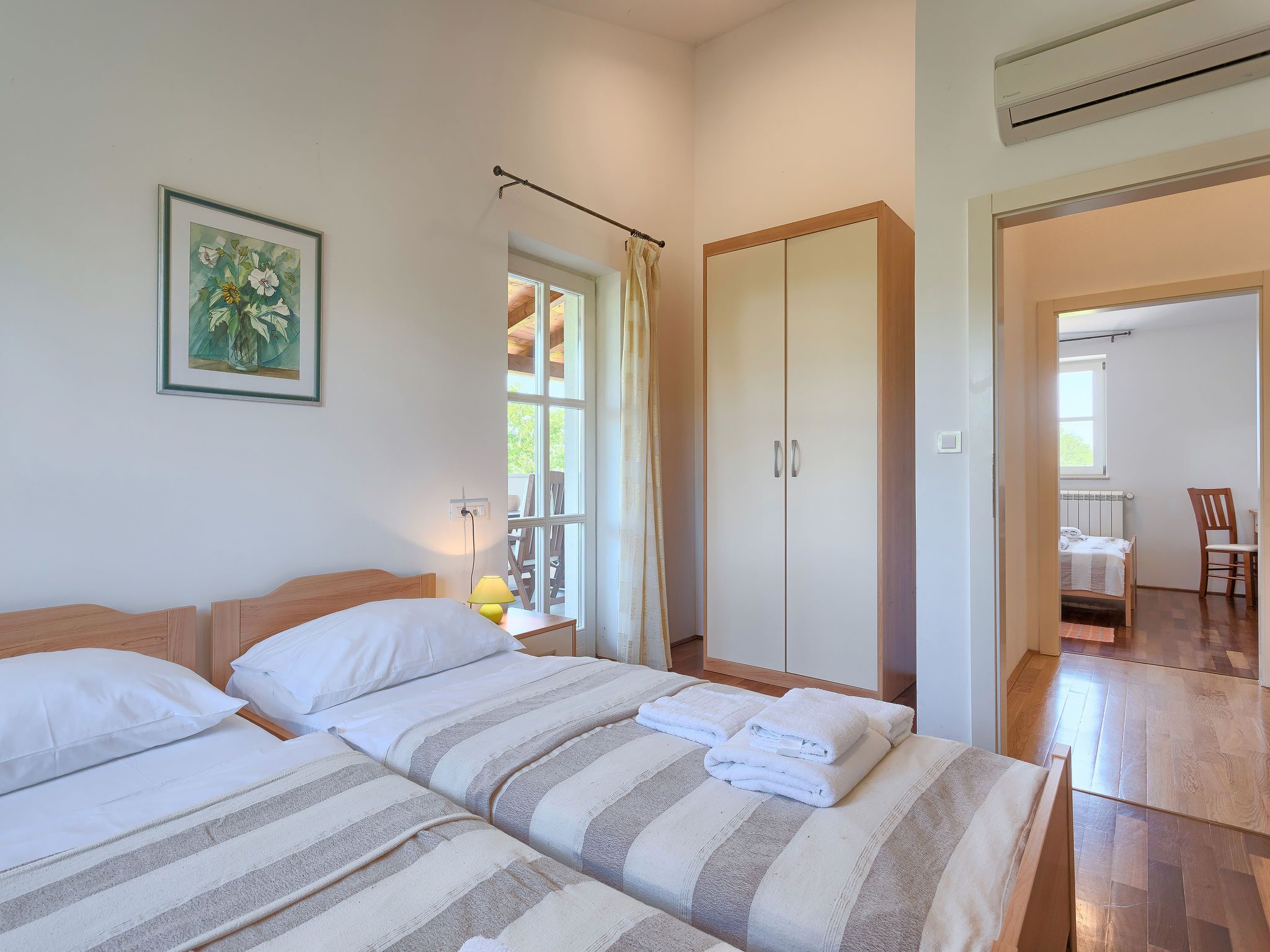 A bedroom with two beds, a wardrobe, a window, and a view of a balcony.