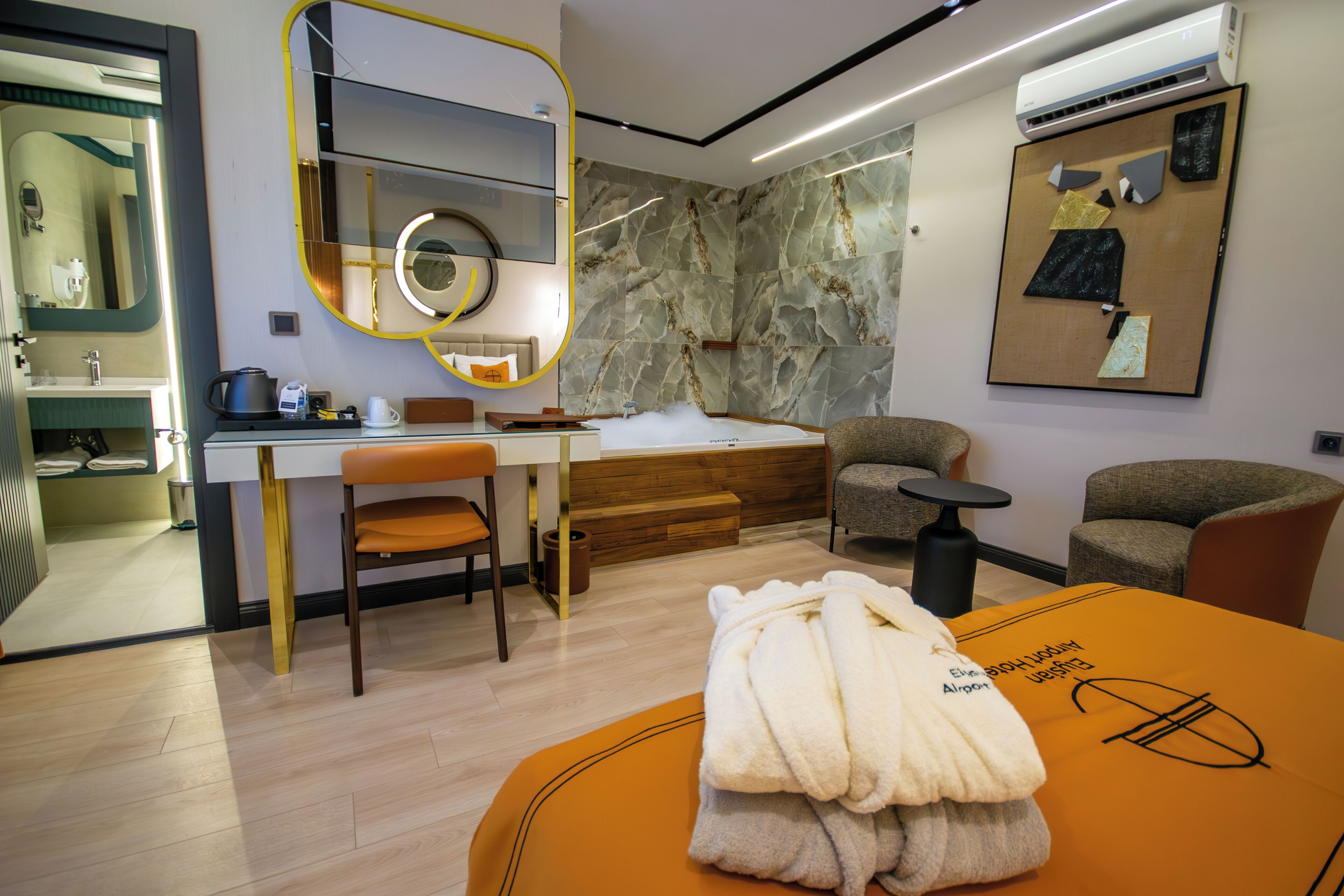 A modern hotel room with a wooden floor, a desk with a chair, a large mirror, a bed with a wooden headboard, and a wall-mounted air conditioning unit.