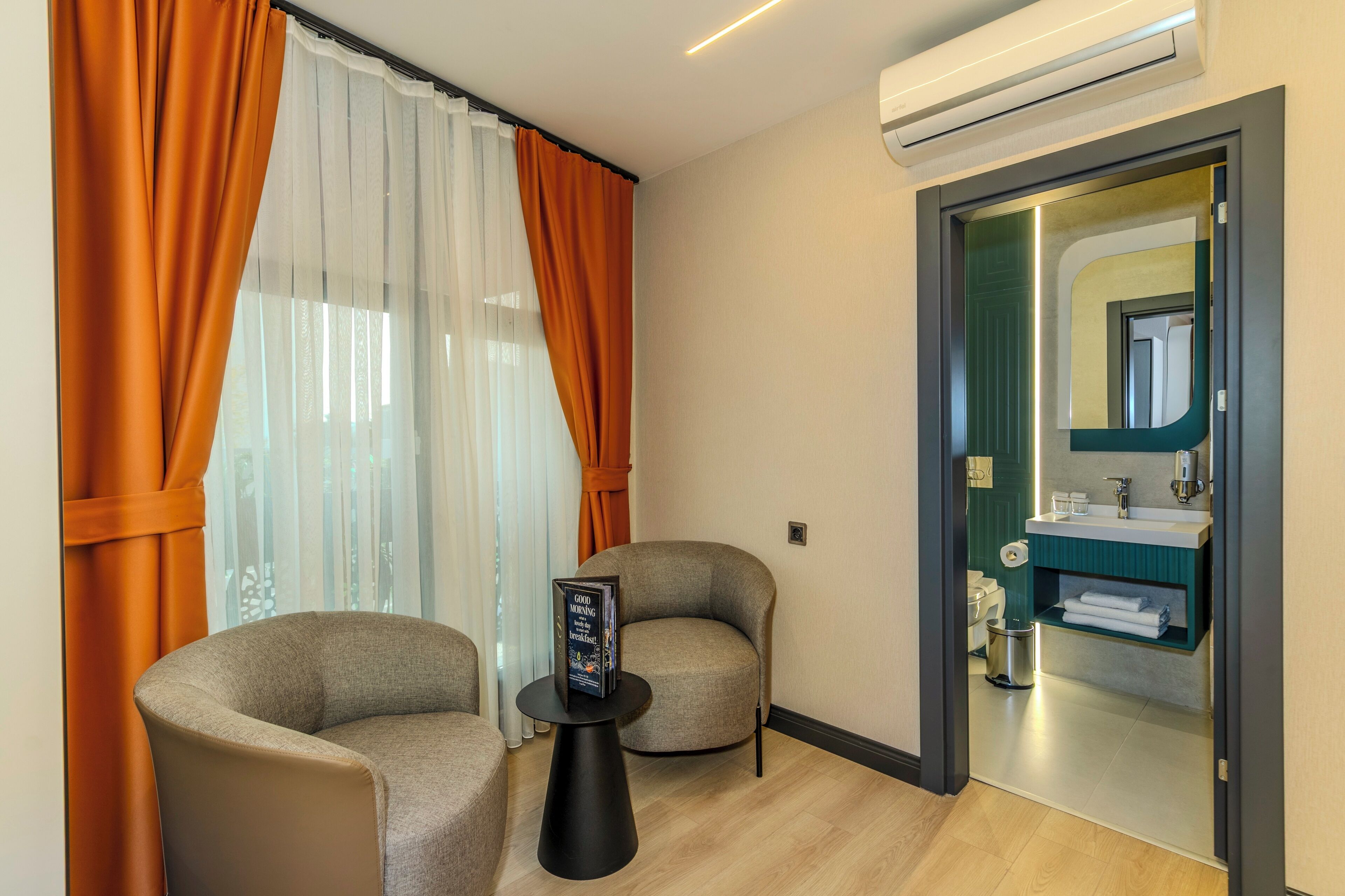 Habitación de hotel moderna con sofá, una mesita con una revista y vista al baño a través de una puerta de vidrio.