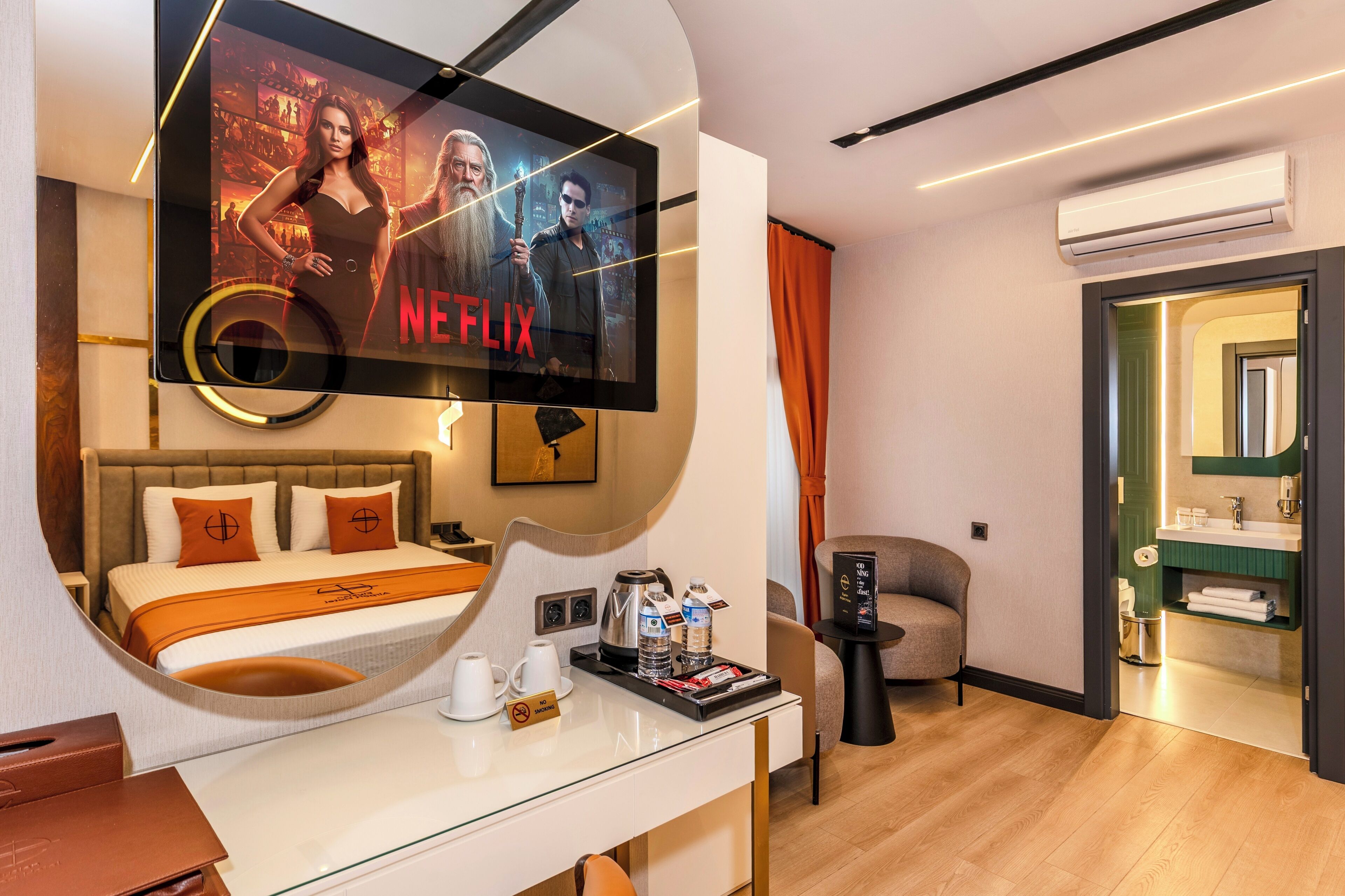 Une chambre d’hôtel dotée d’une grande télévision diffusant une série Netflix, d’un lit avec une tête de lit marron, d’un bureau avec une machine à café et d’une petite chaise.