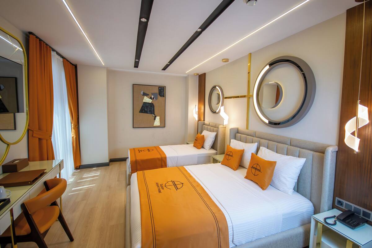 Habitación de hotel con dos camas, un escritorio y una silla. Hay dos mesitas de noche con lámparas, un espejo grande y un cuadro en la pared.