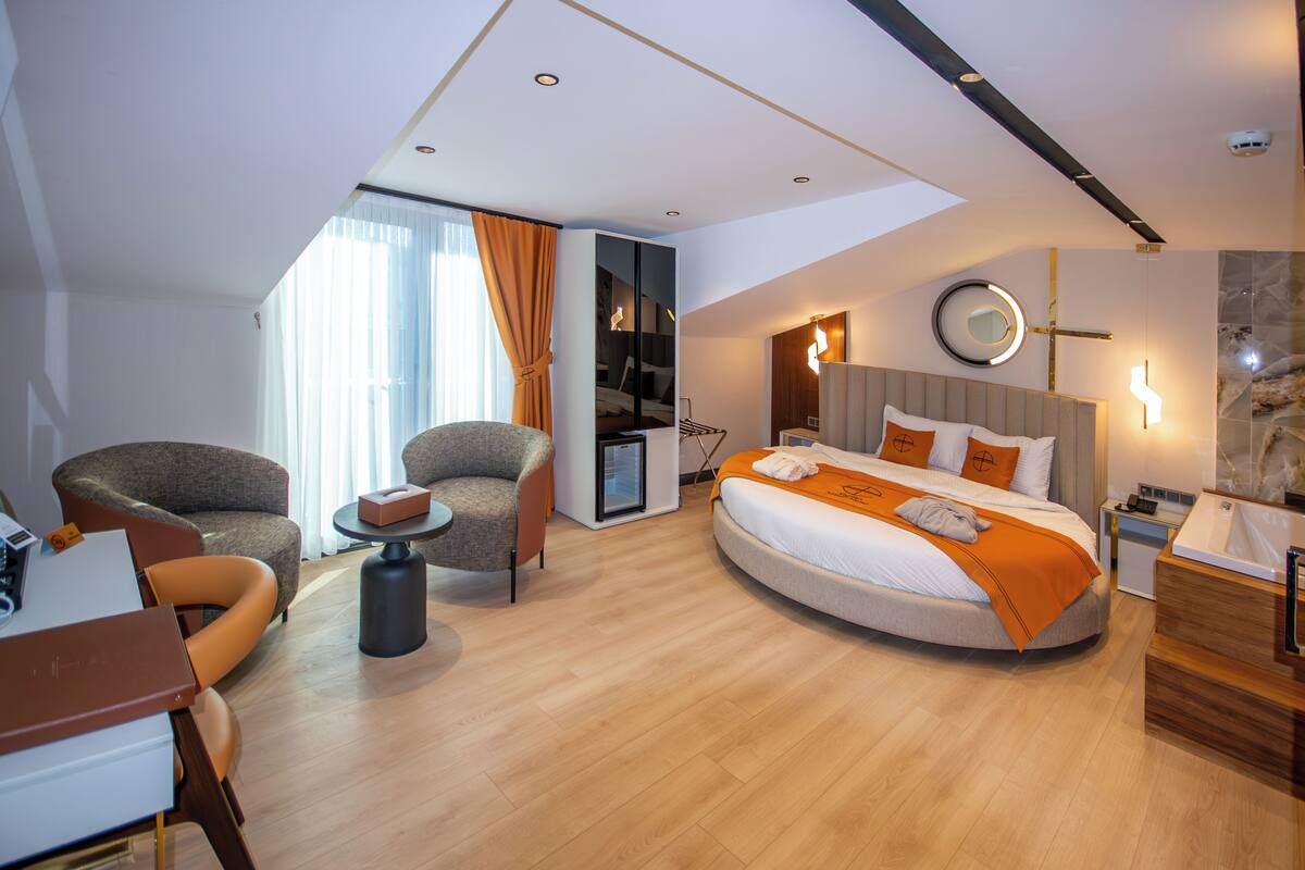 Una habitación de hotel moderna con una cama curva, un área de descanso con dos sillones, una mesita y un escritorio.