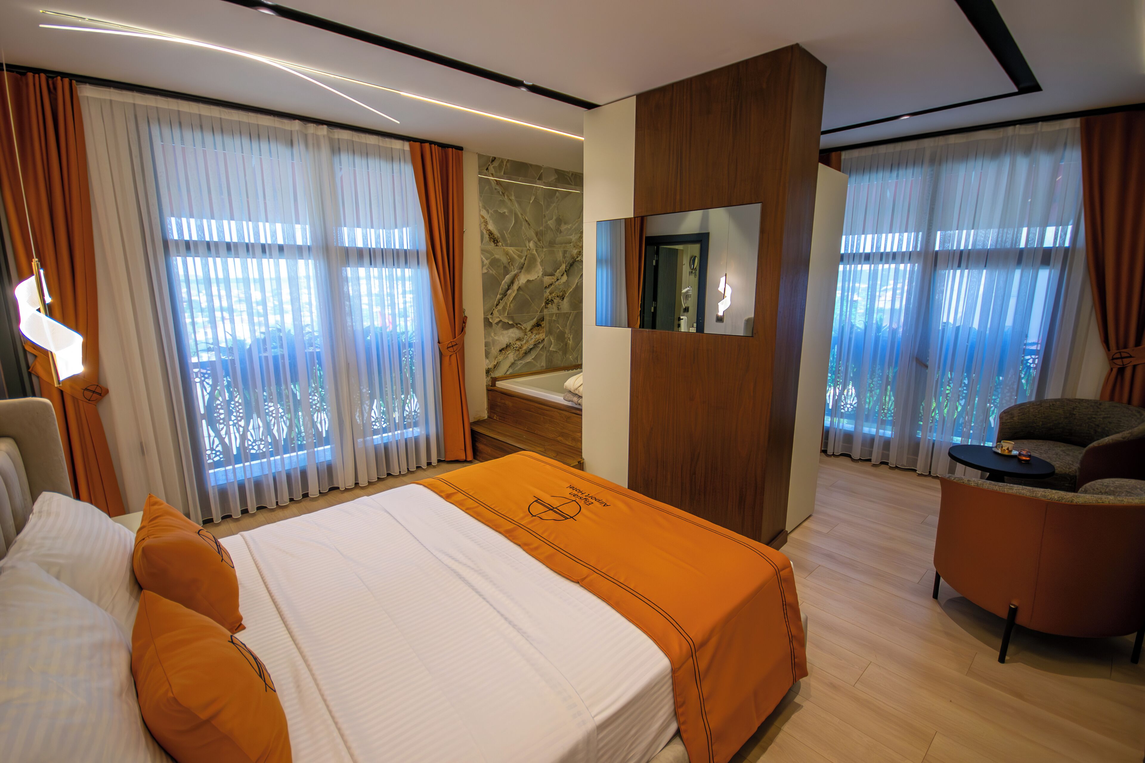 A modern hotel room with a large bed, a sitting area with a sofa and a small table, a bathroom with a mirror, and large windows with curtains.