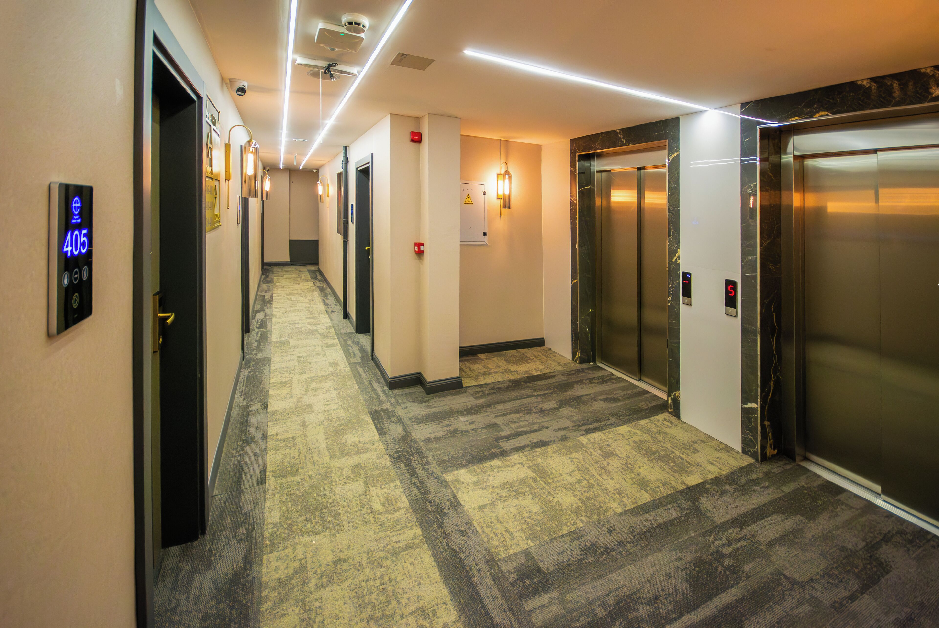 Un pasillo de hotel con piso alfombrado, un ascensor y una pantalla digital que muestra el número de habitación 405.