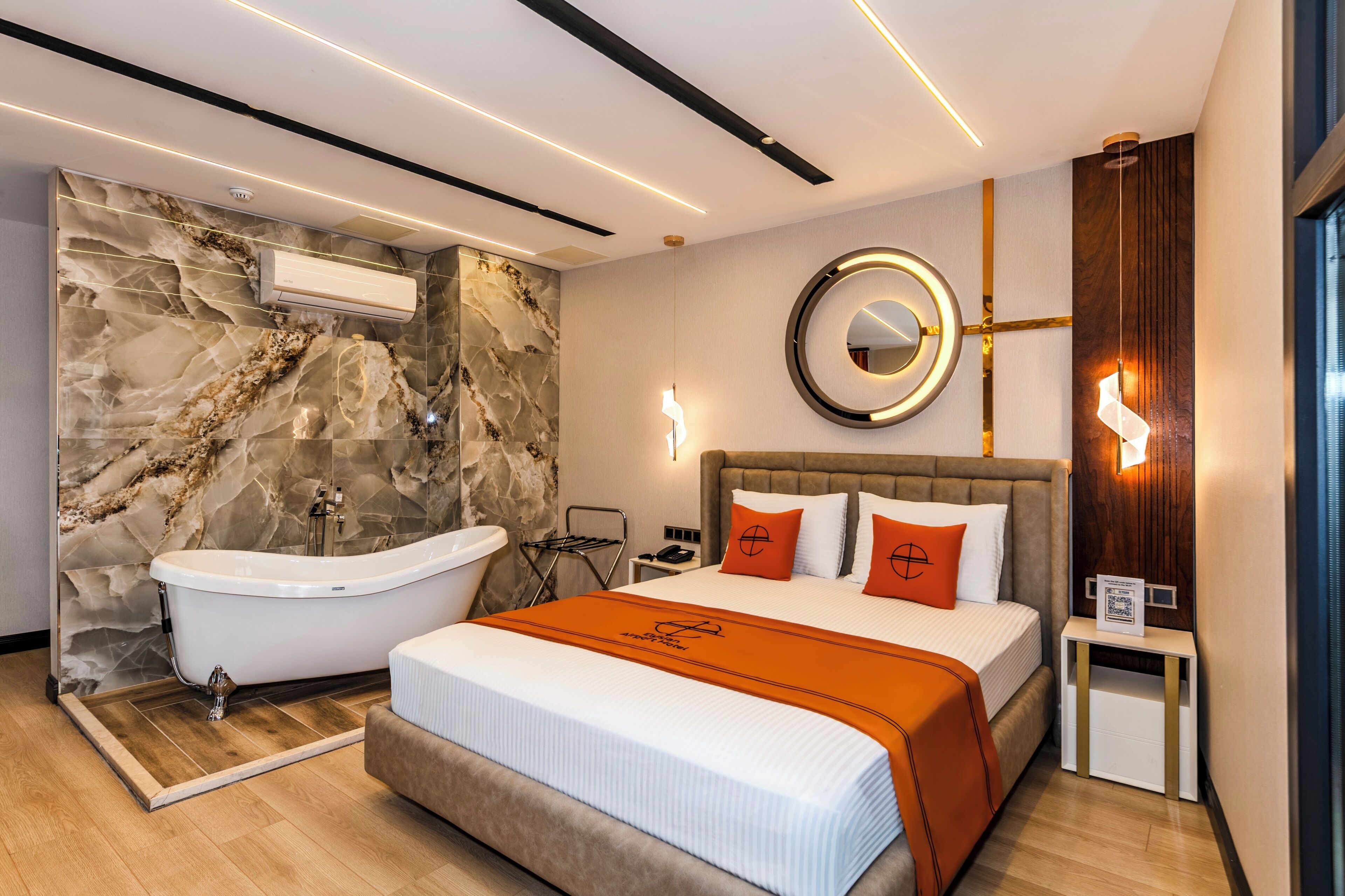 Ein modernes Hotelzimmer mit Marmorwand, einer Badewanne, einem Bett mit orangefarbener Bettwäsche, einem runden Spiegel und einem Nachttisch mit einem QR-Code.
