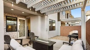 An outdoor patio with a glass-top table, two lounge chairs, a pergola with string lights, a brick wall, and a window.