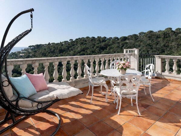 Una terraza con una mesa y sillas blancas, una silla de mimbre con cojines y vistas a una colina boscosa.