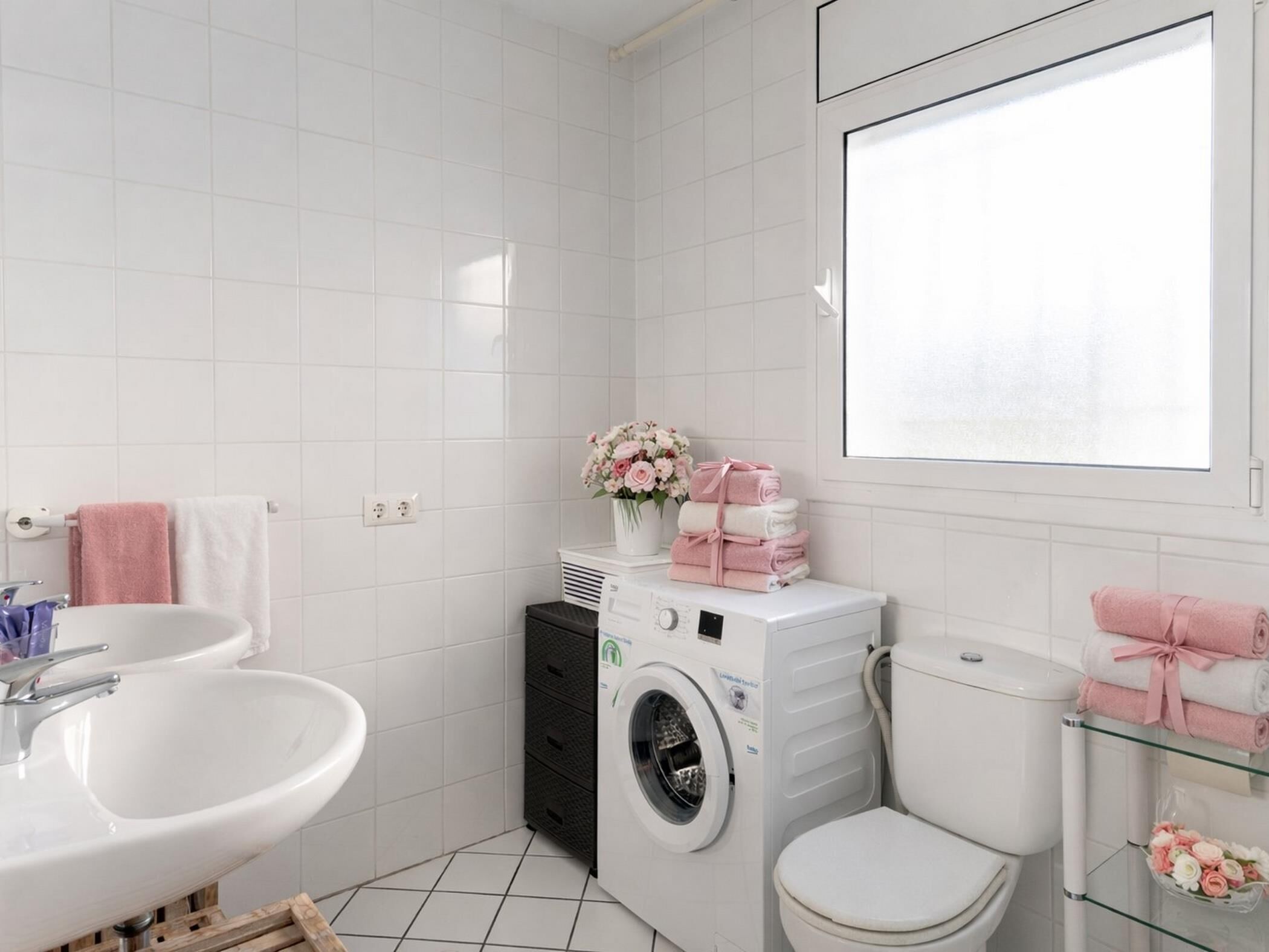 A modern bathroom with white tiles, a sink, a washing machine, a toilet, and a glass shelf with towels.