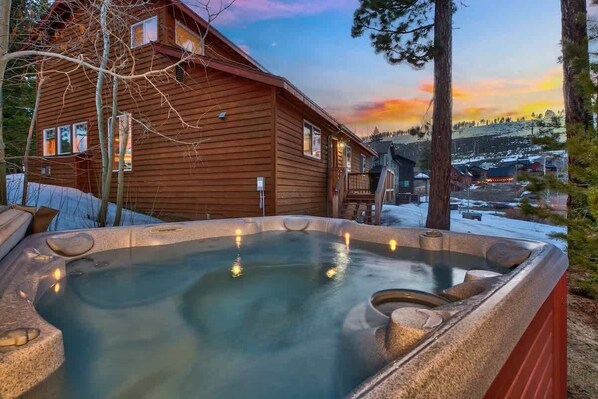 A wooden cabin with a hot tub in front, surrounded by trees and a snowy landscape.