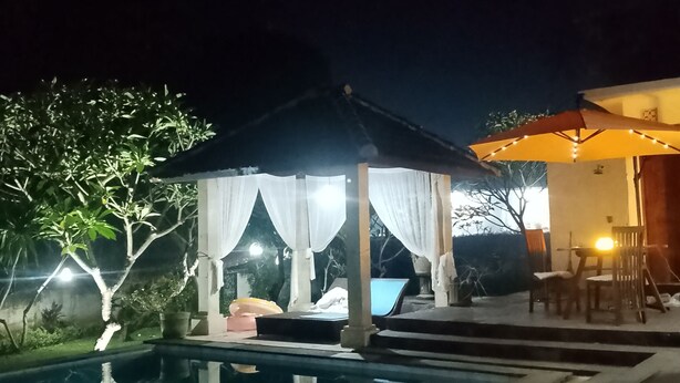 Ein Blick auf einen Poolbereich bei Nacht, mit einem Pavillon, einem Tisch mit Stühlen und einer Laterne.