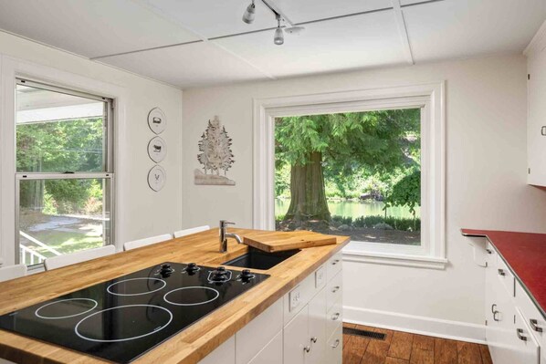 A modern kitchen with a central island, white cabinetry, and a large window offering a view of a tree and greenery.