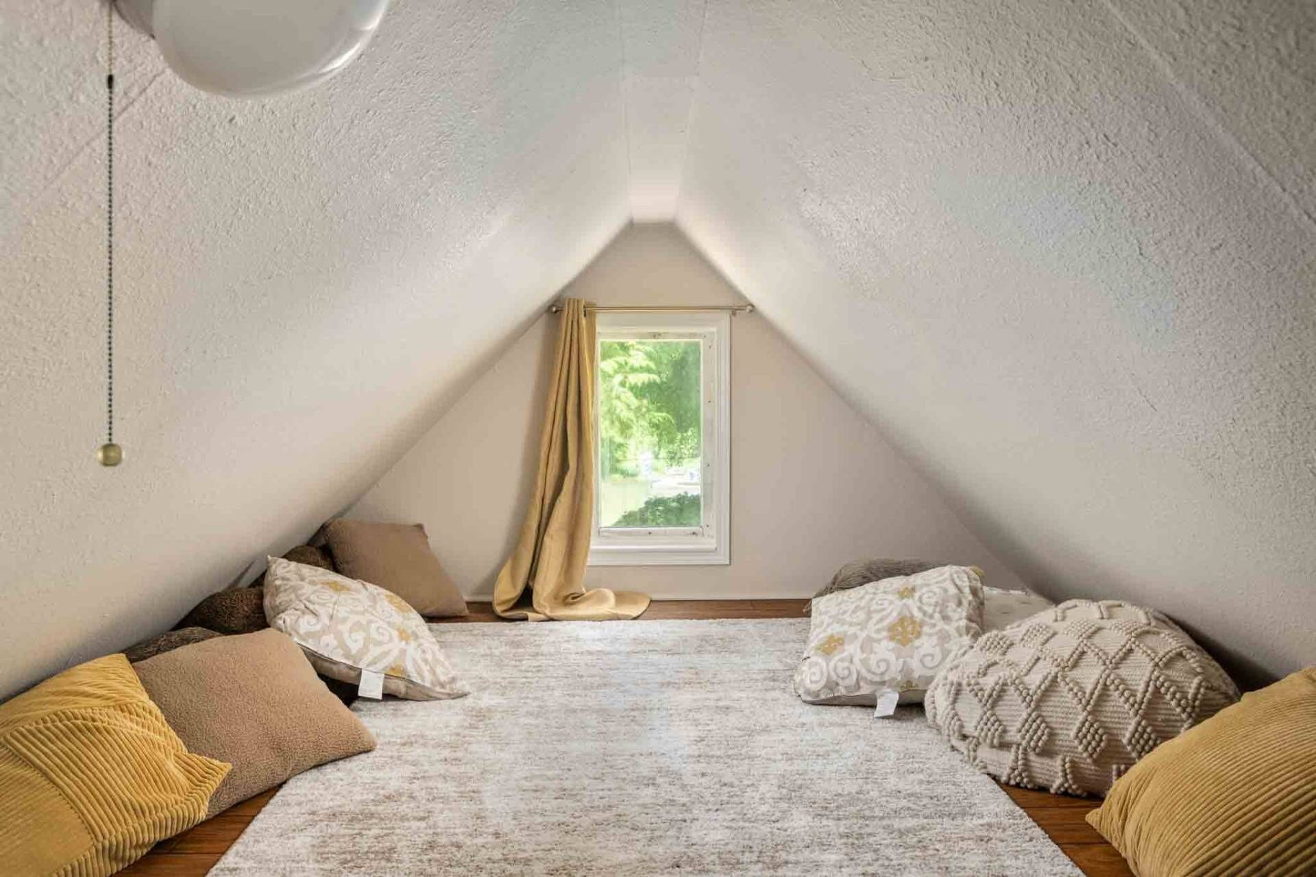 Une chambre mansardée chaleureuse, avec un plafond en pente, une fenêtre donnant sur un paysage verdoyant et une variété d’oreillers aux motifs variés.