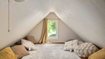 Une chambre mansardée chaleureuse, avec un plafond en pente, une fenêtre donnant sur un paysage verdoyant et une variété d’oreillers aux motifs variés.