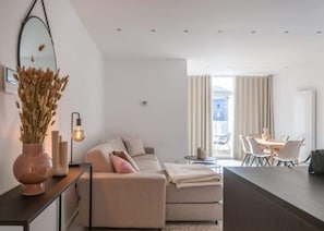 A modern living room with a sofa, a dining table with chairs, a mirror, and a vase with dried flowers.