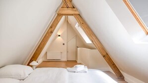 Un dormitorio con techo inclinado, vigas de madera, una cama, una mesita de noche, una lámpara y un radiador.