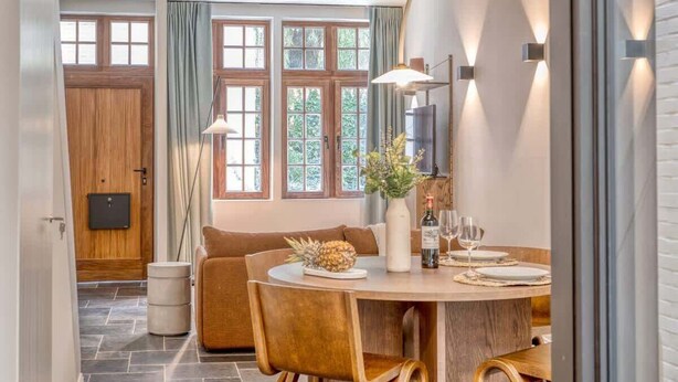Una cocina moderna con vigas de madera a la vista, suelo de baldosas de piedra, una zona de comedor con mesa redonda y sillas de madera, y ventanas grandes con cortinas.