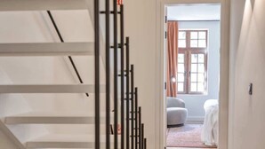 Un interior moderno con una escalera de madera, una puerta visible que da a una habitación con cama y cortinas, y un suelo de color claro.