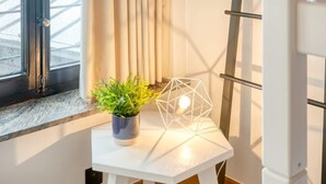 Une pièce avec une fenêtre, un rideau, une petite table avec une plante et un luminaire au design géométrique, ainsi qu’un radiateur blanc.