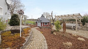 Een pad van stenen loopt naar een klein, rood-wit gebouw met een puntig dak, omgeven door een tuin met mulch en beplanting, en een houten pergola.