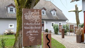 Een rustiek bord met tekst in een vreemde taal, een boom en een huis met pannendak op de achtergrond.