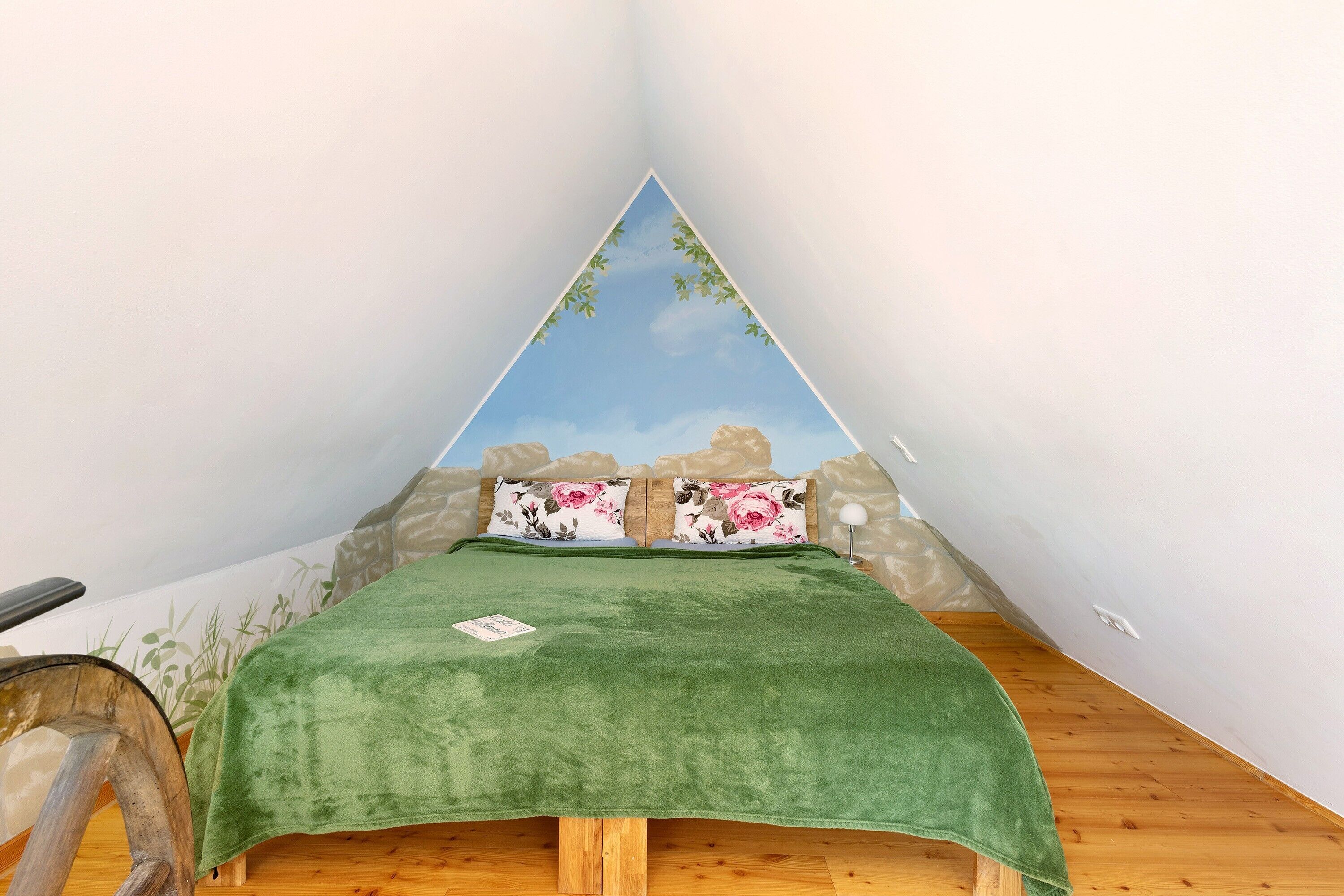 A triangular room with a green bedspread, floral pillows, and a view of rocks and sky through a triangular window.