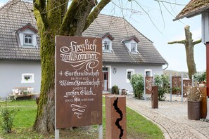 Un cartello rustico con testo in lingua straniera, un albero e, sullo sfondo, una casa con tetto in tegole.