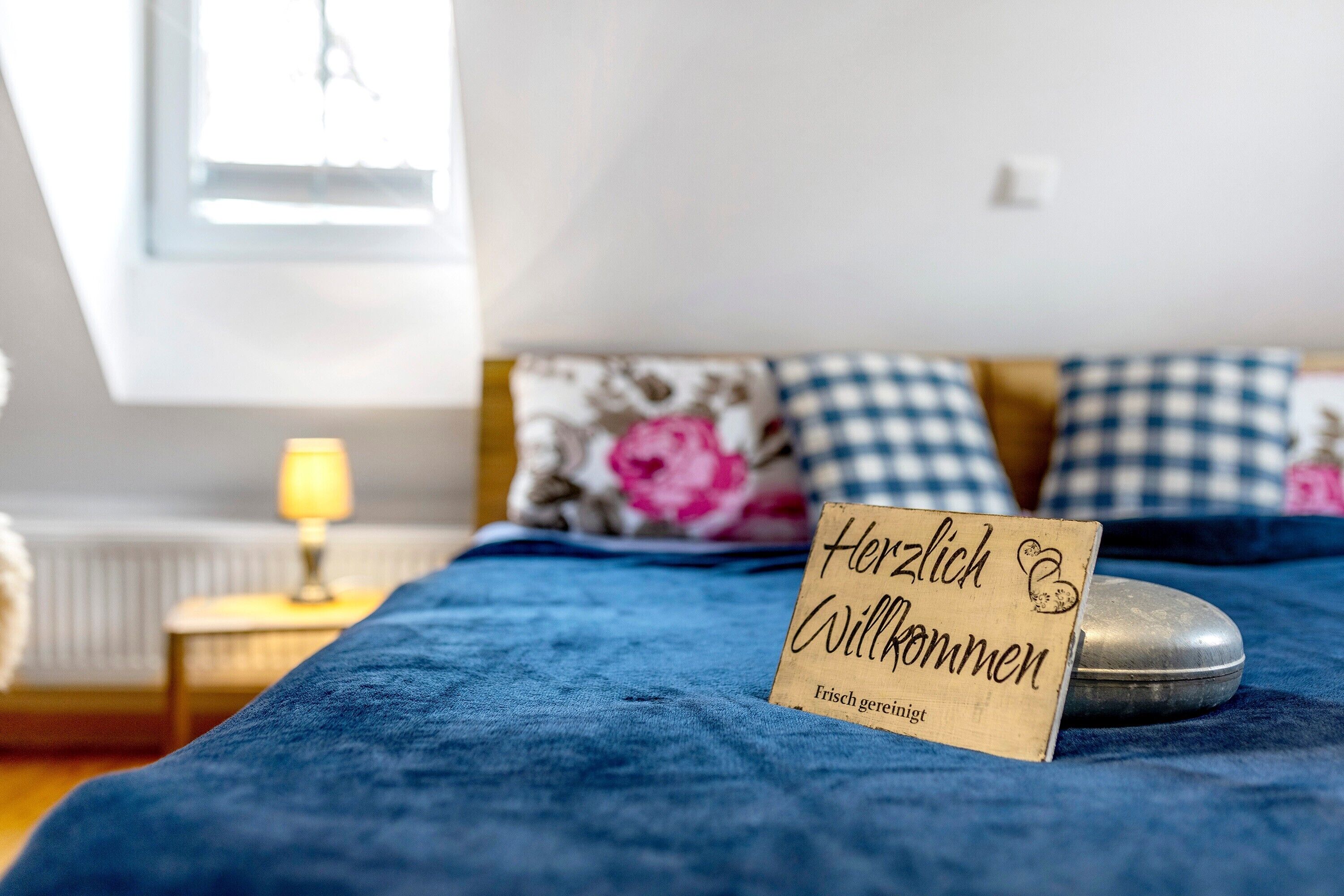 Un letto con una coperta blu, una lampada da comodino e un cartello di legno con la scritta 'Herzlich Willkommen'.