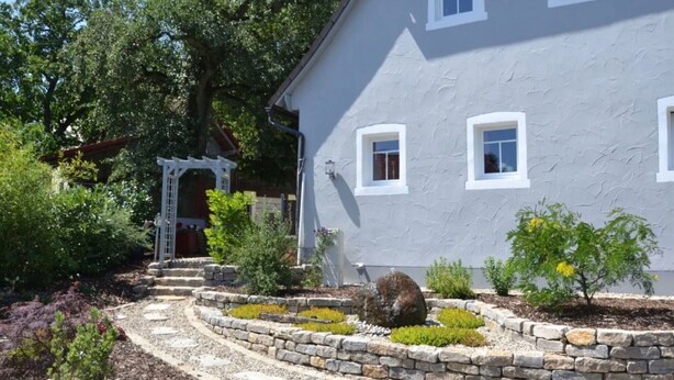 A house with a stone pathway leading to a white door, surrounded by various plants and a garden. - Apartment 'Stub'n Rettl' with Shared Terrace, Shared Garden and Wi-Fi (Waischenfeld)