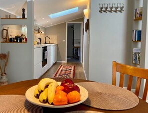 Une cuisine moderne avec des meubles blancs, un coin repas avec une table en bois et une assiette de fruits, et un couloir menant à une chambre.