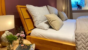 Un lit avec des coussins à motifs, une table de chevet avec un livre et des fleurs, et une fenêtre donnant sur des arbres.