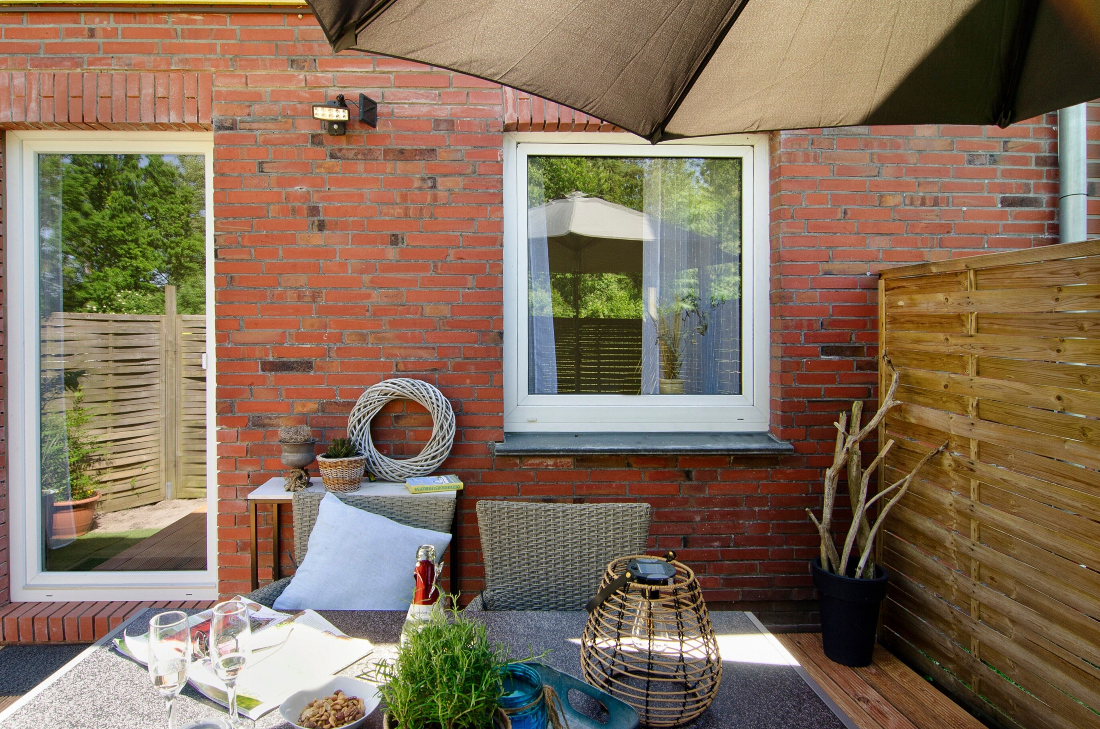 Une terrasse avec une table dressée pour deux personnes, un panier en osier, une plante en pot et une fenêtre donnant sur un paysage verdoyant.