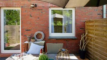 Une terrasse avec une table dressée pour deux personnes, un panier en osier, une plante en pot et une fenêtre donnant sur un paysage verdoyant.
