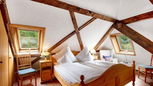 Une chambre avec un lit en bois, deux tables de chevet, une chaise et un bureau avec une lampe. Deux fenêtres laissent entrer la lumière naturelle.