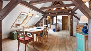 Une chambre mansardée avec des poutres apparentes, une table à manger avec des chaises, une fenêtre et une vue sur la cuisine adjacente.