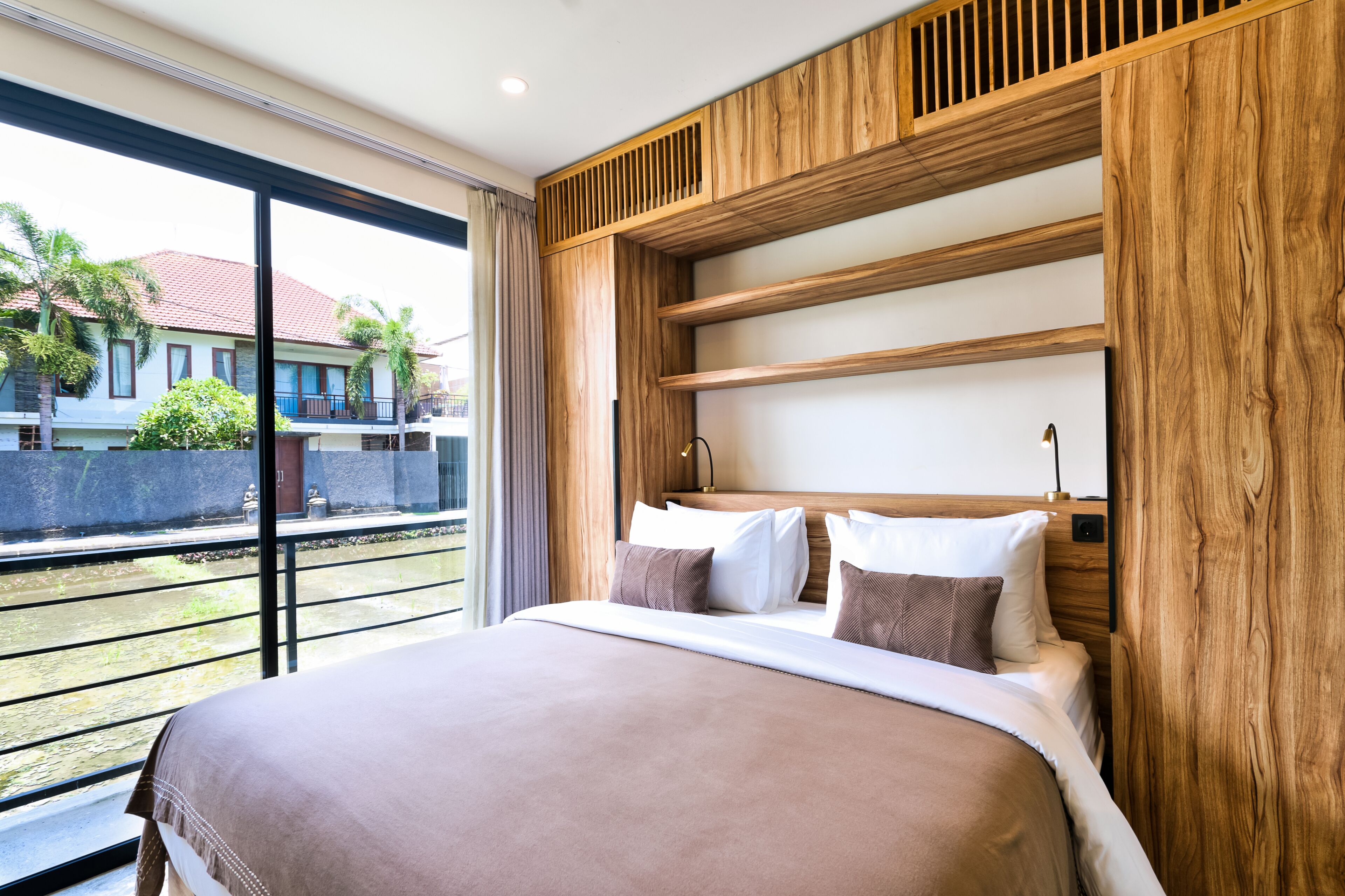 A modern hotel room with a large bed, wooden headboard, and a view of a building and palm trees outside.