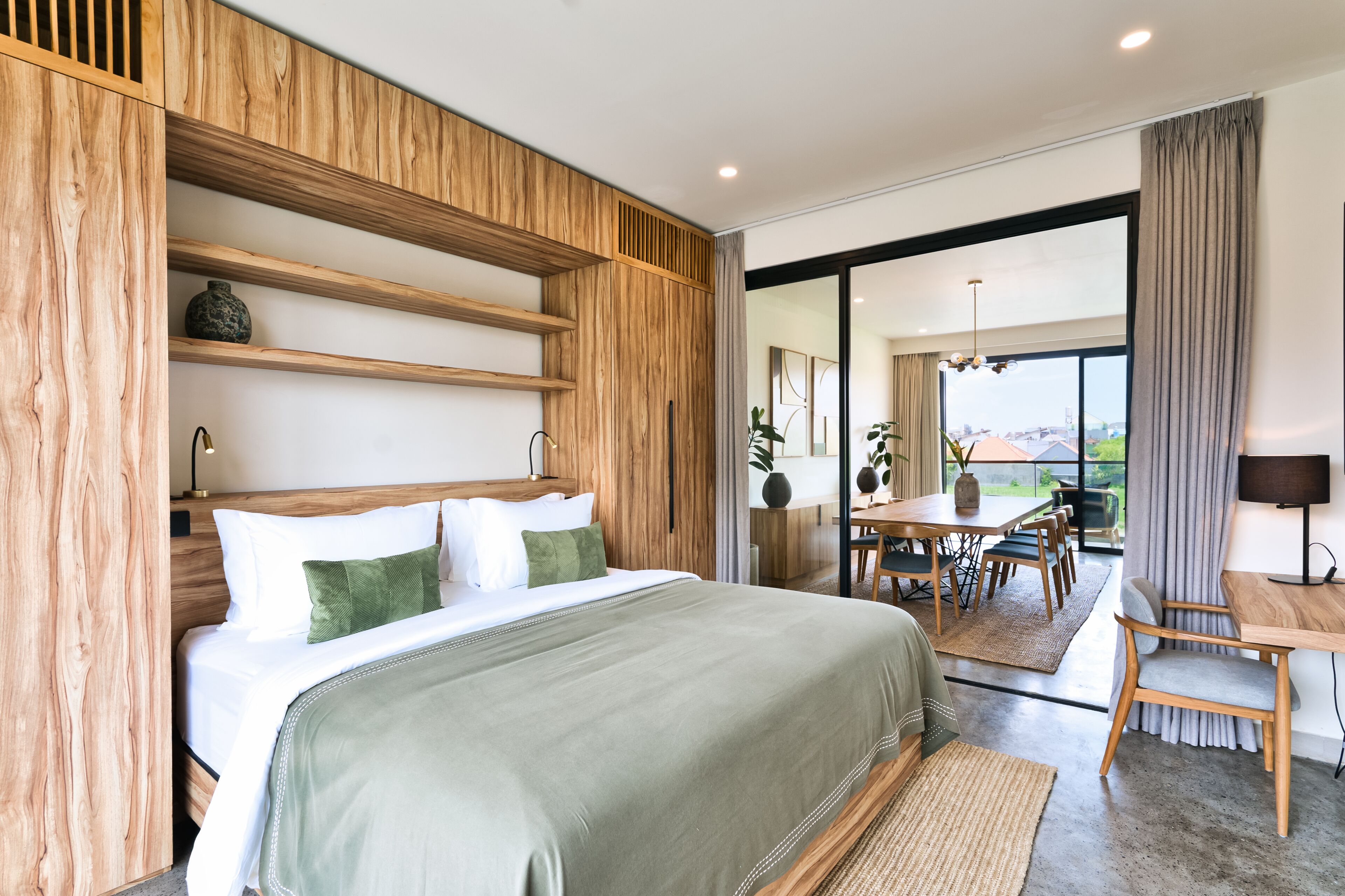 A modern hotel room with a large bed, wooden paneling, a dining area, and a view through the glass door.