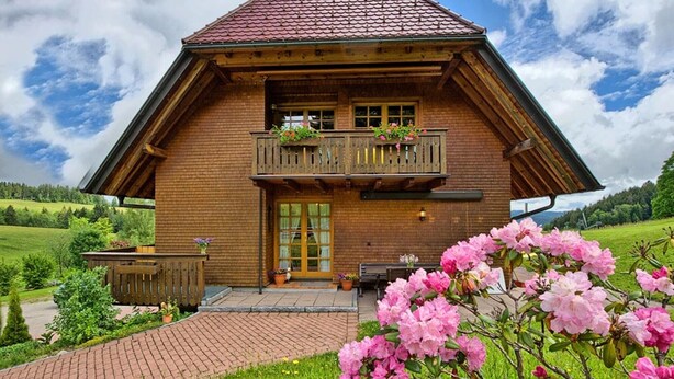 Un chalet en bois avec un toit en pente, un balcon agrément é de plantes en pot et un chemin en briques menant à l’entrée.