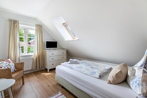 Un dormitorio con cama, televisión, ventana con cortinas, una silla y una mesita.
