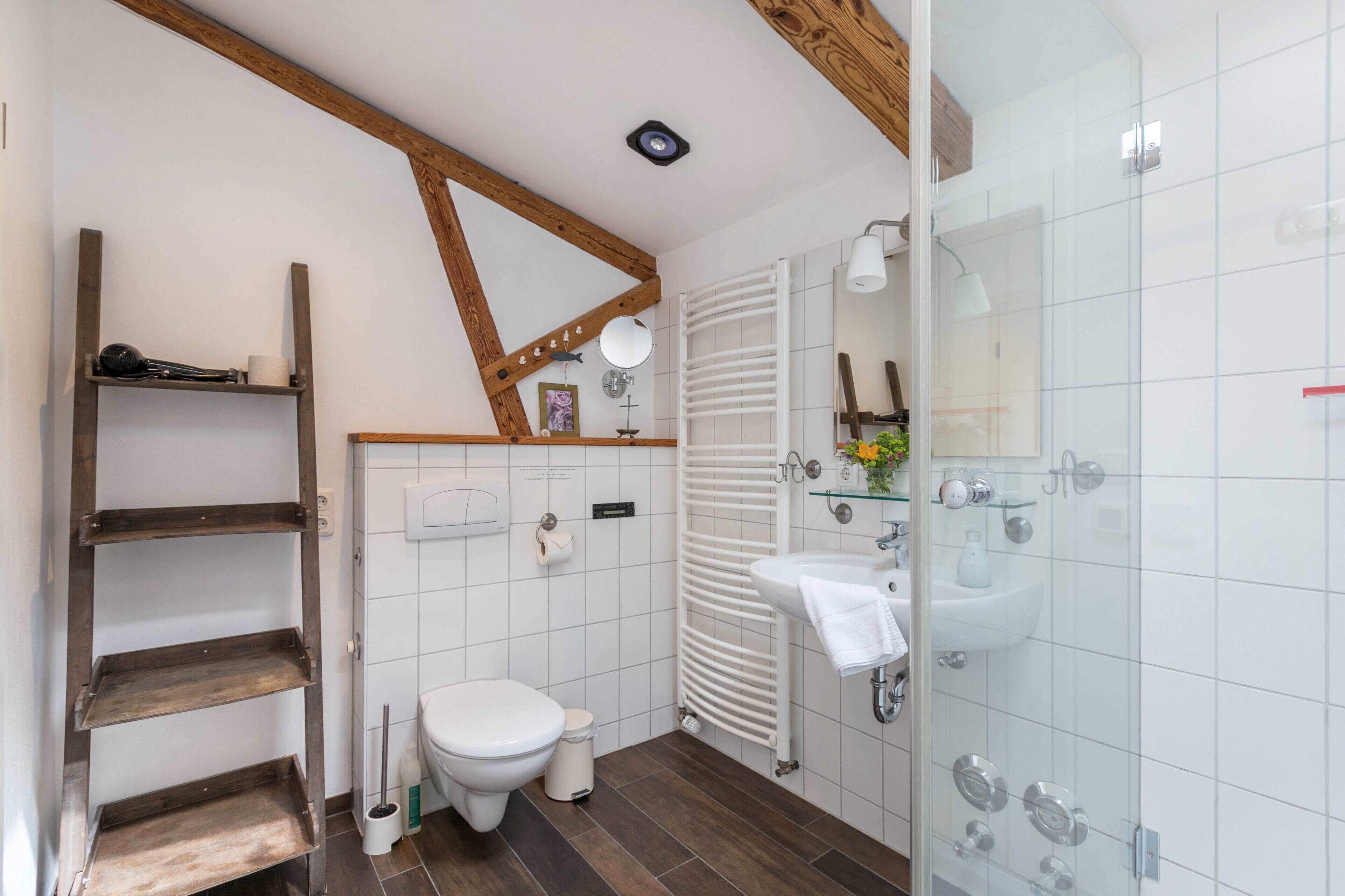 Un baño con inodoro, lavamanos, cabina de ducha de vidrio, estante de madera y pared de azulejos blancos.