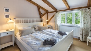 A bedroom with a wooden ceiling, a white bed with floral bedding, a nightstand, a chair, and a window with curtains. - Holiday Apartment "Alter Heuboden" with Private Garden, Panoramic Views and Wi-Fi (Steinbergkirche)
