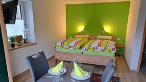 Una habitación pequeña y moderna con una cama, una mesa de comedor con sillas y un televisor colgado en la pared.