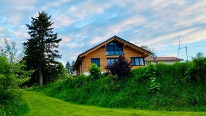 A two-story wooden house with large windows, surrounded by lush greenery and a clear sky.