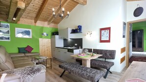 A cozy room with a wooden ceiling, a dining table with benches, a sofa, and a kitchen area.