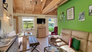 A cozy room with a wooden ceiling, a sofa, a bench, a TV, a window, and a green wall with framed pictures.
