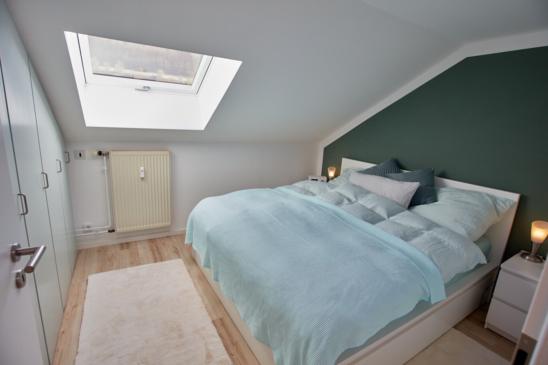 Quarto com claraboia, cama com várias almofadas, abajures, radiador e porta com puxador.