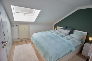 Een slaapkamer met dakraam, een bed met veel kussens, nachtlampjes, een radiator en een deur met een handgreep.