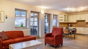 Una sala de estar con sofá, sillón y mesa de centro, una cocina con armarios y comedor, y ventanas amplias con vistas.