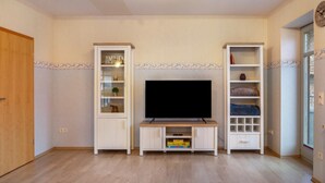 Una habitación con una televisión sobre un soporte de madera, dos armarios blancos con puertas de vidrio y una ventana con un reloj.